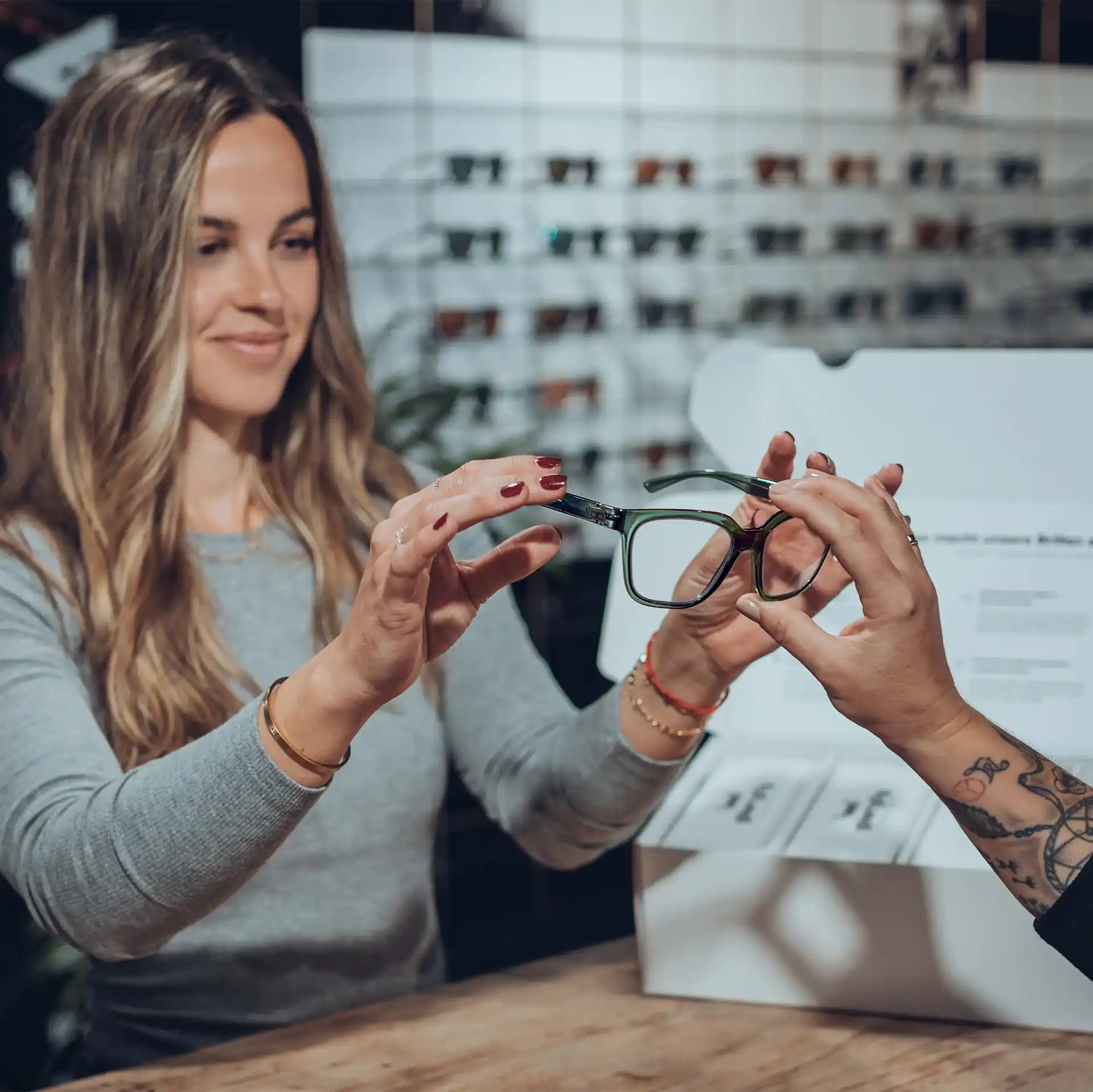 Eine Frau mit langen, welligen Haaren hält eine Brille in den Händen und betrachtet sie genau. Eine weitere Hand, mit Tattoos verziert, reicht ihr die Brille. Im Hintergrund sind weitere Brillenmodelle an einer Wand ausgestellt.