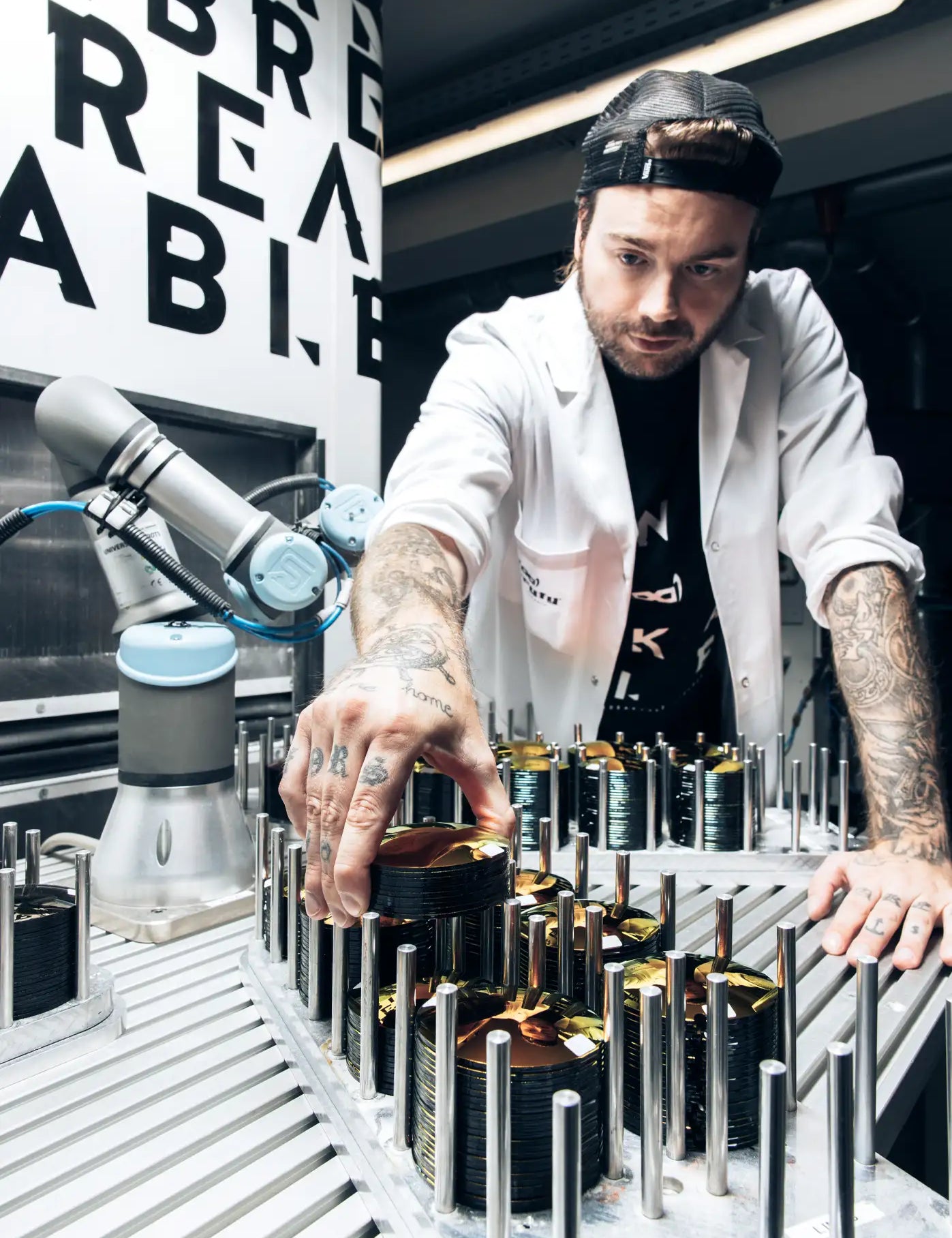 Ein tätowierter Techniker in weißem Laborkittel und schwarzer Cap arbeitet konzentriert in einer High-Tech-Fertigungsanlage. Er stapelt runde, gold-beschichtete Gläser auf einer Metallhalterung. Im Hintergrund ist ein kollaborativer Roboterarm zu sehen, der Teil des automatisierten Produktionsprozesses ist.