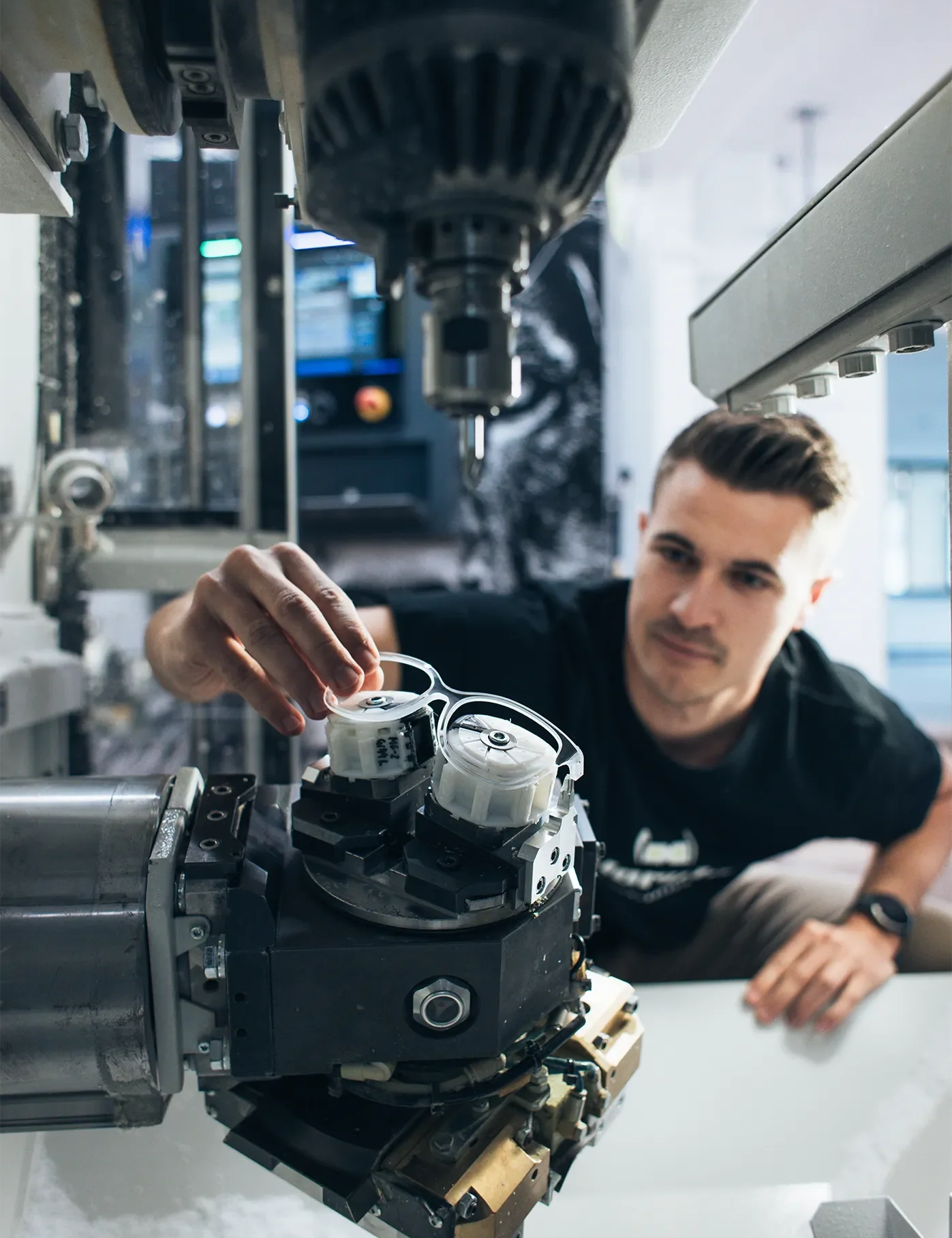 Ein junger Mann mit dunklem T-Shirt und Smartwatch arbeitet an einer hochpräzisen CNC-Maschine. Er justiert vorsichtig ein Bauteil in der Maschine, während im Hintergrund ein Bildschirm mit technischen Daten zu sehen ist. Die Szene zeigt moderne Fertigungstechnologie in einer professionellen Werkstatt.