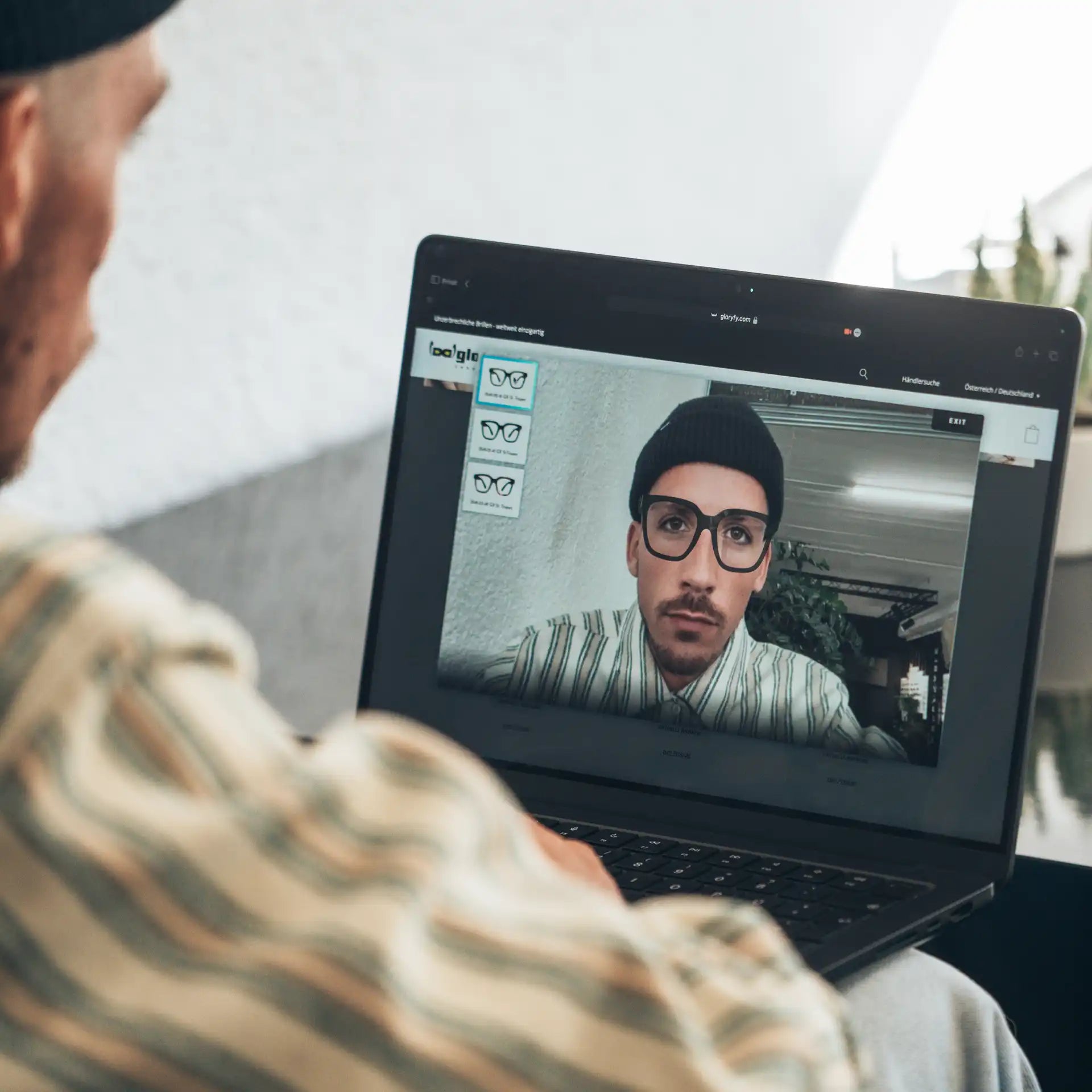 Ein Mann sitzt mit einem Laptop auf dem Schoß und probiert virtuell verschiedene Brillenmodelle mit einer Online-Anprobe. Auf dem Bildschirm ist sein Gesicht mit einer digitalen Brille zu sehen. Die Szene zeigt modernes Online-Shopping.