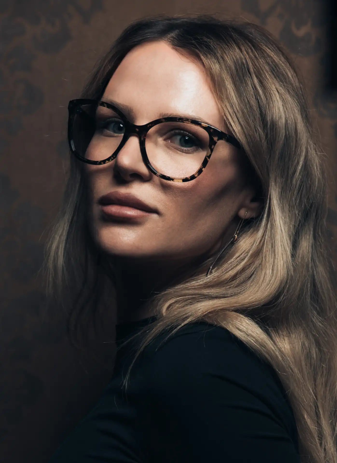 Eine Frau mit langen, blonden Haaren trägt eine große, runde Brille mit einem dezenten Muster im Rahmen. Sie blickt leicht zur Seite mit einem ausdrucksstarken Blick. Der Hintergrund ist dunkel und wirkt edel, passend zu ihrem eleganten Look mit dezentem Make-up und langen Ohrringen.