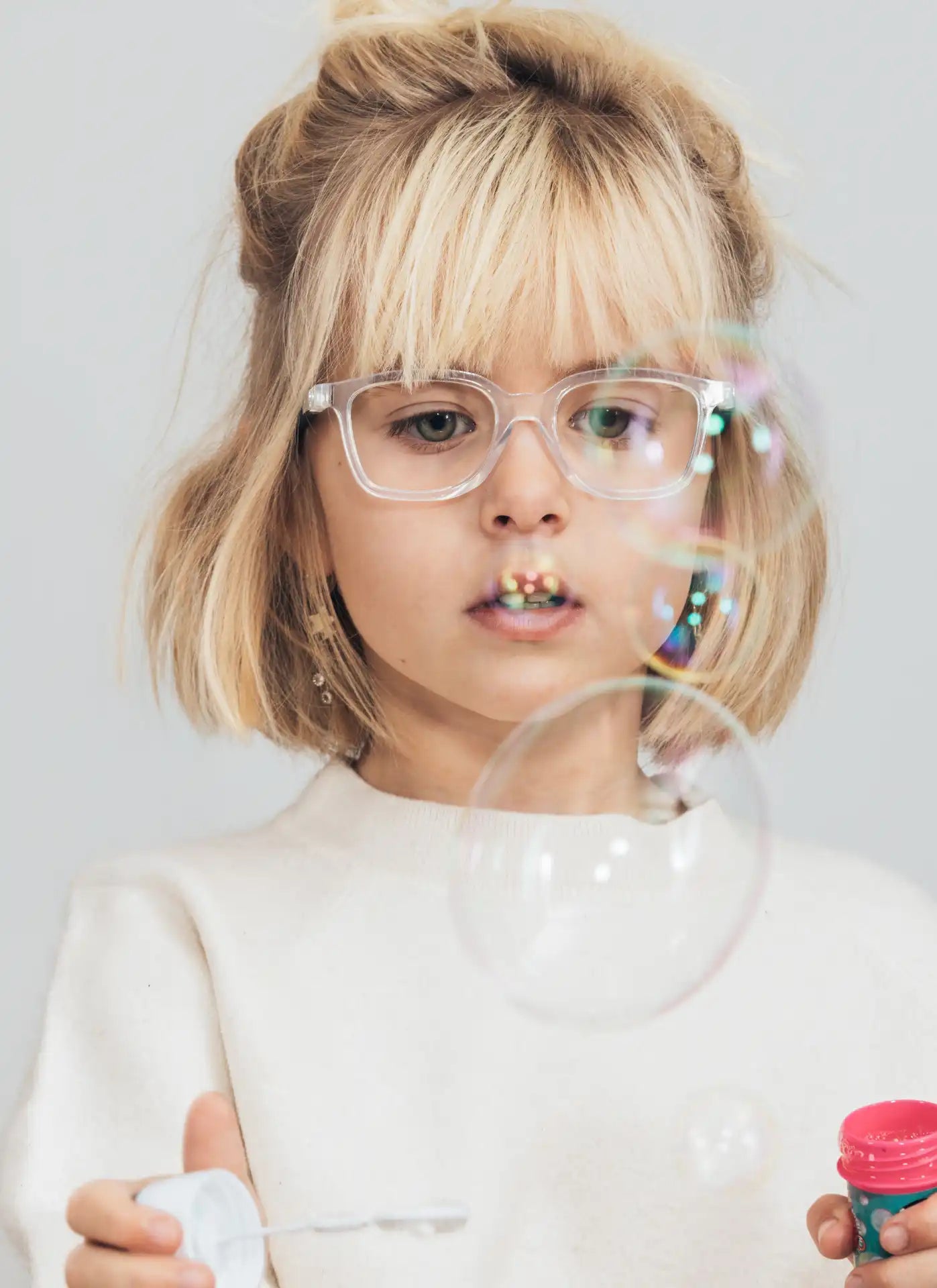 Ein blondes Mädchen mit einem verspielten Haardutt trägt eine durchsichtige Brille und bläst konzentriert Seifenblasen. Ihr Gesichtsausdruck zeigt Staunen, während die schimmernden Blasen vor ihr in der Luft schweben. Sie trägt einen hellen Pullover, und der Hintergrund ist minimalistisch und hell.