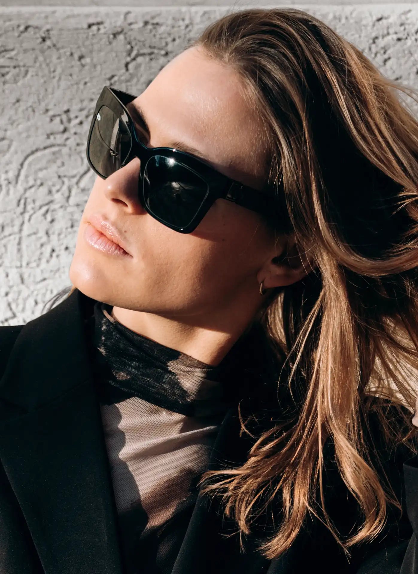 Eine elegante Frau mit langen, leicht gewellten Haaren trägt eine große, schwarze Sonnenbrille. Sie lehnt sich mit entspannter Haltung gegen eine helle Wand, während Sonnenlicht auf ihr Gesicht fällt. Ihr Outfit kombiniert einen schwarzen Blazer mit einem transparenten, gemusterten Oberteil.