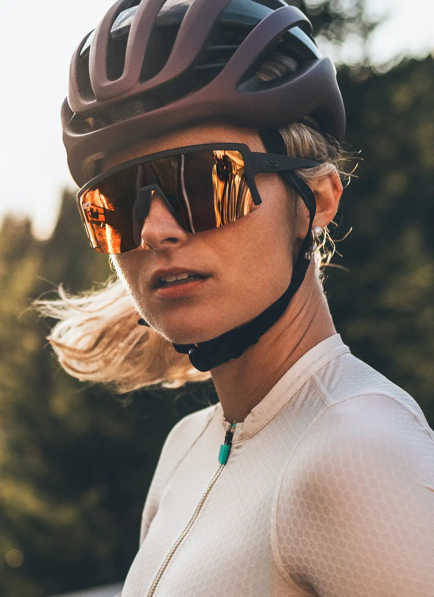 Eine sportliche Frau trägt einen Fahrradhelm und eine große, verspiegelte Sport-Sonnenbrille mit rötlich getönten Gläsern. Ihr blondes Haar weht leicht im Wind, während sie konzentriert zur Seite blickt. Der Hintergrund zeigt eine natürliche Umgebung mit Bäumen im warmen Sonnenlicht.