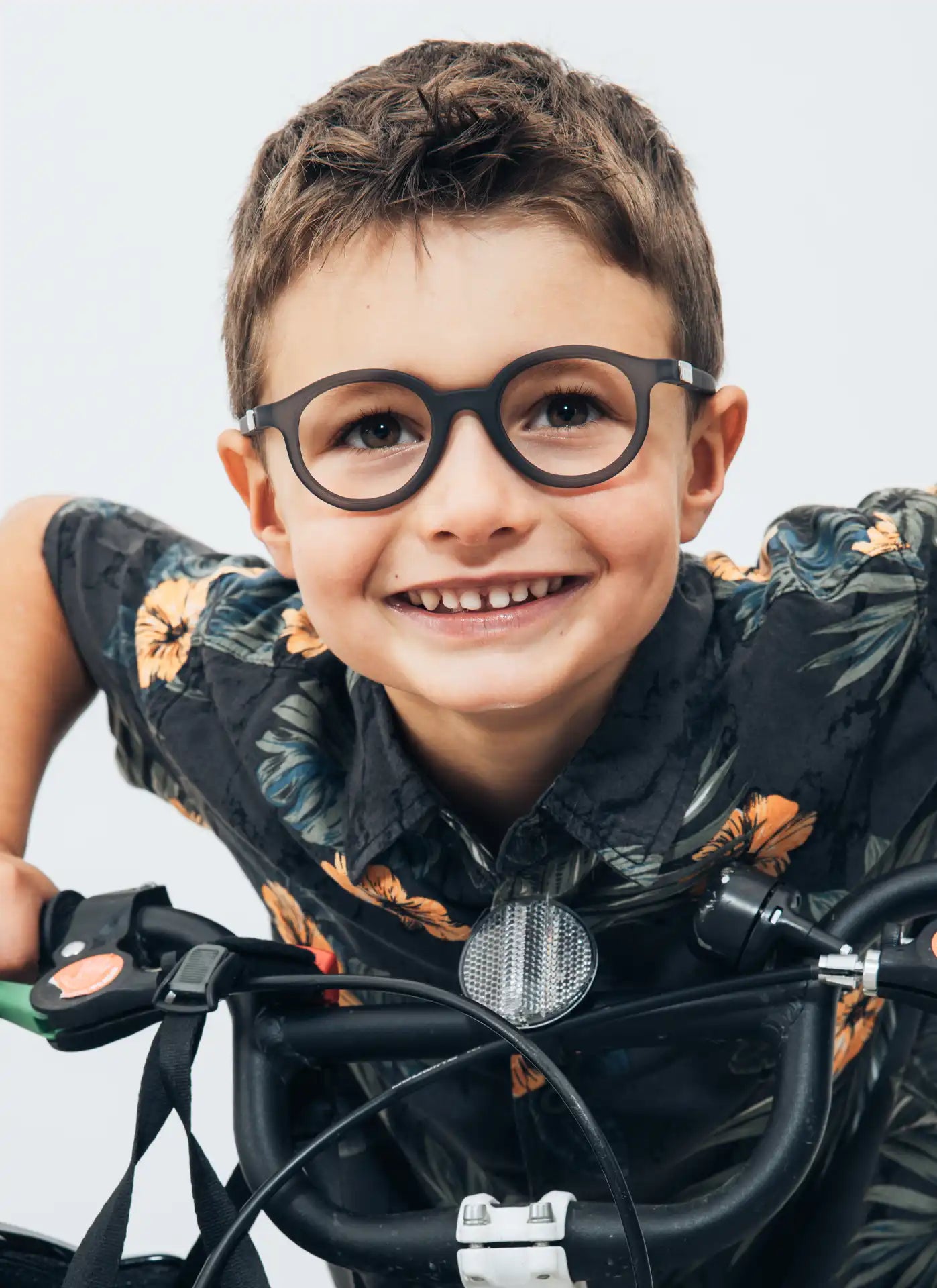 Ein fröhlicher Junge mit braunen Haaren und runder Brille lehnt sich über den Lenker seines Fahrrads. Er trägt ein bunt gemustertes Hemd und strahlt mit einem breiten Lächeln. Der Hintergrund ist minimalistisch und hell.