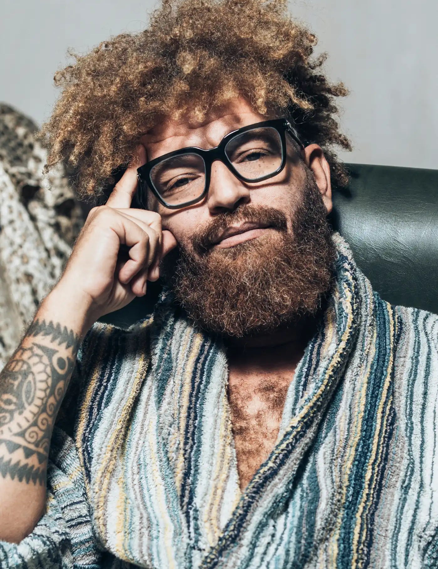 Ein Mann mit lockigem, vollem Haar und Bart trägt eine große, schwarze Brille und einen gemütlichen, gestreiften Bademantel. Er sitzt entspannt auf einem Sessel, stützt seinen Kopf mit der Hand ab und blickt mit einem nachdenklichen Ausdruck in die Kamera.