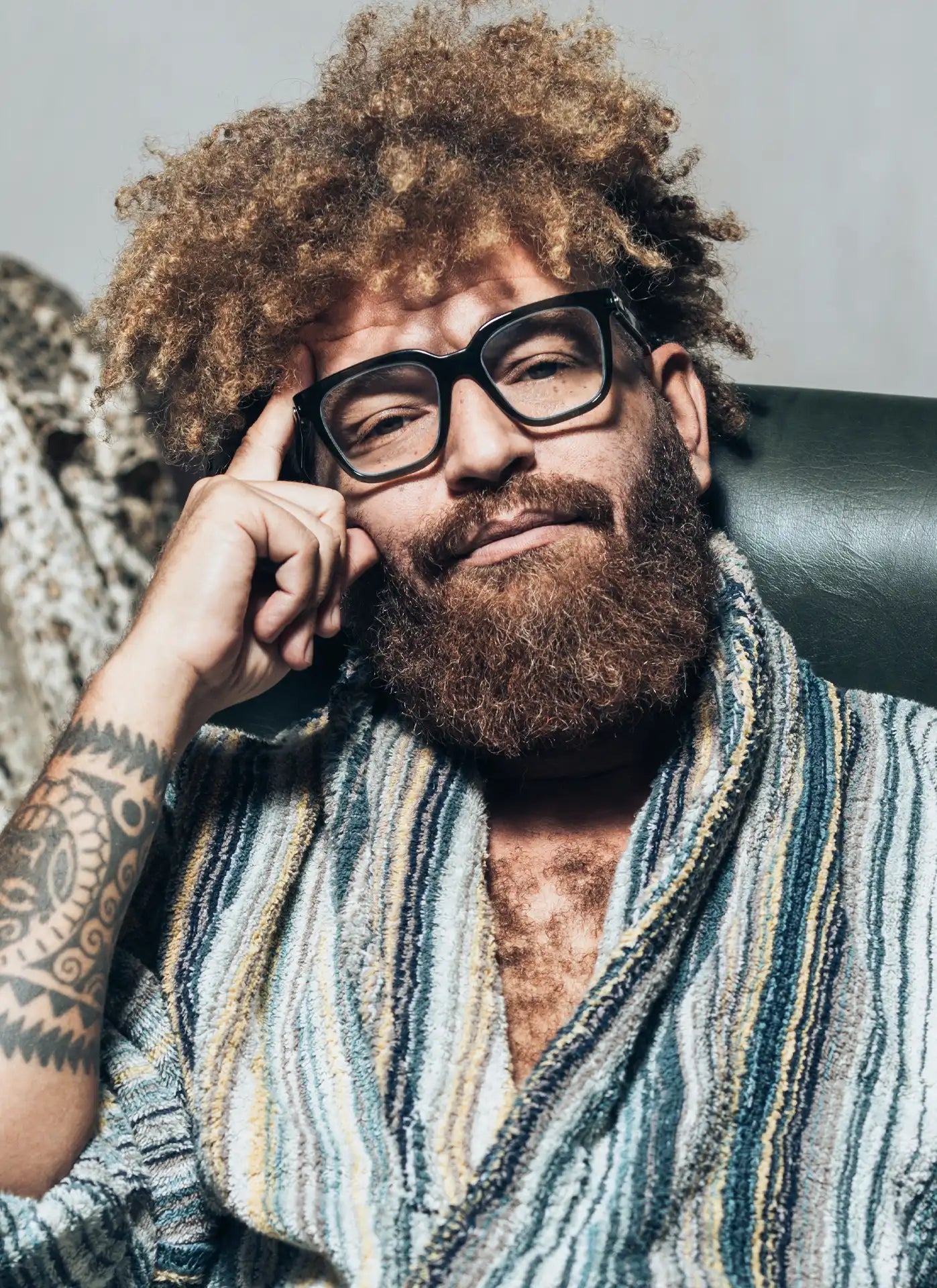 Ein Mann mit lockigem, vollem Haar und Bart trägt eine große, schwarze Brille und einen gemütlichen, gestreiften Bademantel. Er sitzt entspannt auf einem Sessel, stützt seinen Kopf mit der Hand ab und blickt mit einem nachdenklichen Ausdruck in die Kamera.