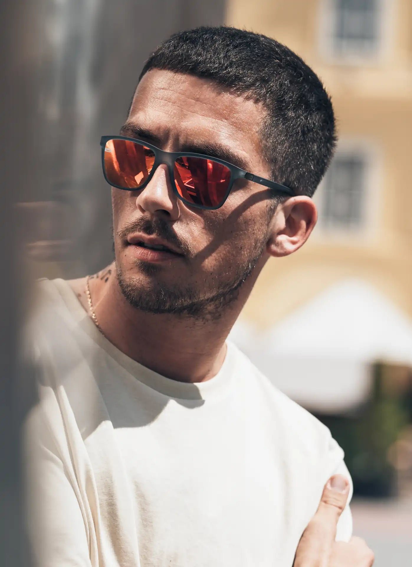 Ein Mann mit kurzem Haar und leichtem Bart trägt eine Sonnenbrille mit roten, verspiegelten Gläsern und ein helles T-Shirt. Er lehnt lässig gegen eine Wand und blickt zur Seite. Der sonnige Hintergrund zeigt eine mediterrane Stadtarchitektur.