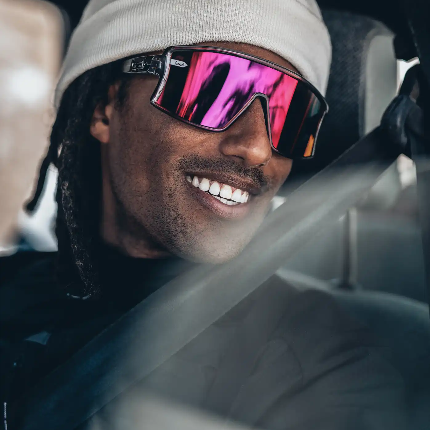 Ein Mann mit weißer Beanie und verspiegelter Sport-Sonnenbrille mit pinken Gläsern sitzt im Auto und lächelt breit. Er trägt einen Sicherheitsgurt, und das Licht spiegelt sich auf der Fensterscheibe. Die Szene wirkt entspannt und urban.