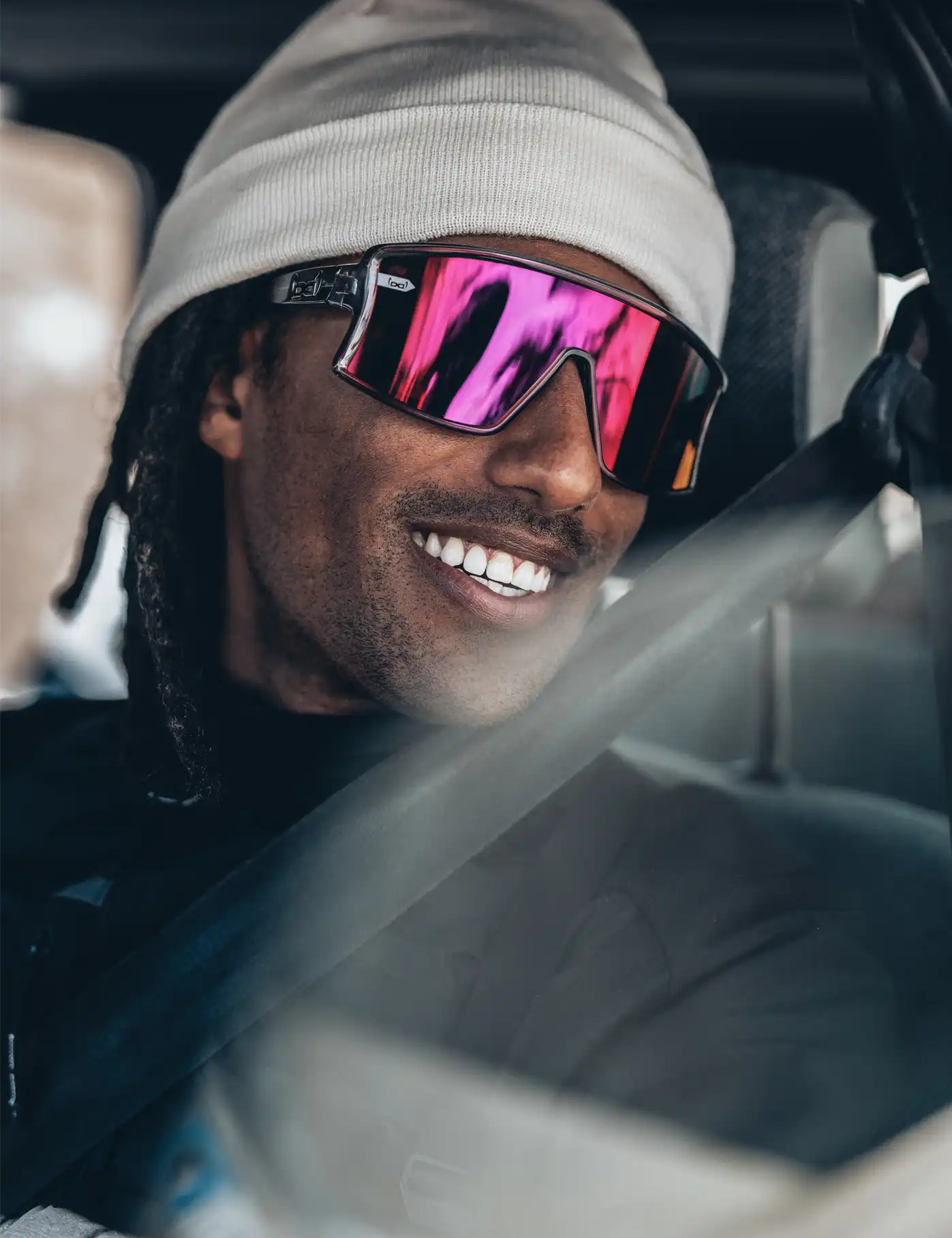 Ein Mann mit weißer Beanie und verspiegelter Sport-Sonnenbrille mit pinken Gläsern sitzt im Auto und lächelt breit. Er trägt einen Sicherheitsgurt, und das Licht spiegelt sich auf der Fensterscheibe. Die Szene wirkt entspannt und urban.