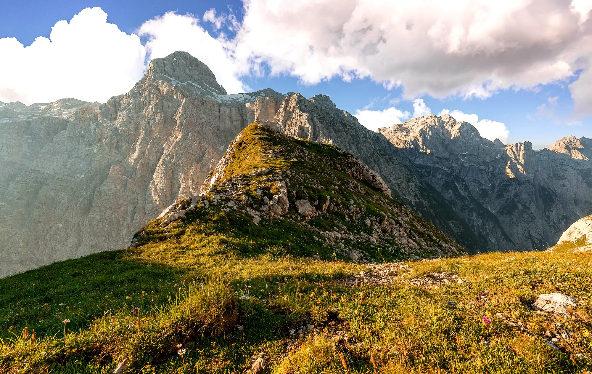 Berglandschaft in warmem Abendlicht: Im Vordergrund eine grüne, mit Felsen durchsetzte Wiese, dahinter ragt ein markanter Felsgipfel steil empor. Im Hintergrund sind weitere graue Gebirgsketten unter blauem Himmel mit weißen Wolken zu sehen.